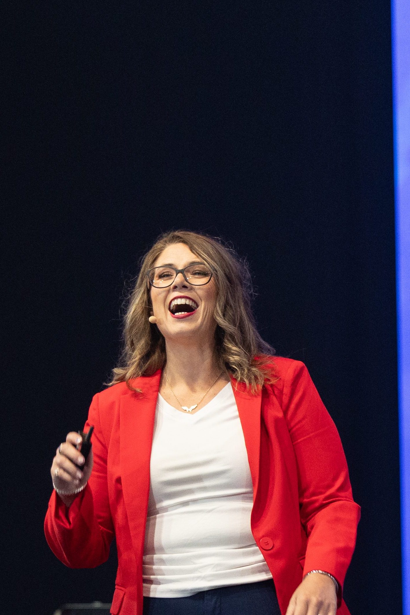 A woman in a red blazer and white shirt smiling while speaking on stage, holding a microphone.