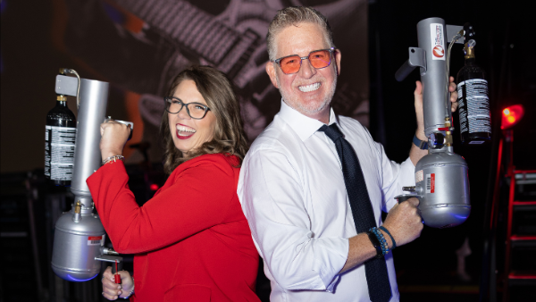 Two people pose back to back, each holding a fire extinguisher. One is a woman in a red blazer with glasses, and the other is a man in a white shirt and tie. The background features stage lighting and a graphic of a guitar.